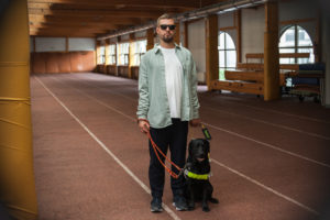 Młody mężczyzna w ciemnych okularach stoi na bieżni w hali sportowej wraz z psem przewodnikiem.