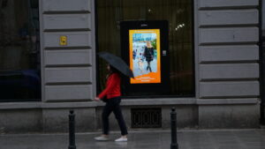 Przed ekranem wyświetlającym przekaz Fundacji Vis Maior przechodzi pani z parasolką